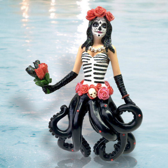 The Catrina Ornament by December Diamonds features a skeletal face with a corset adorned in roses and a small skull, complemented by black octopus tentacles. It holds red roses and wears a rose headdress, all hand-painted against a light blue background.