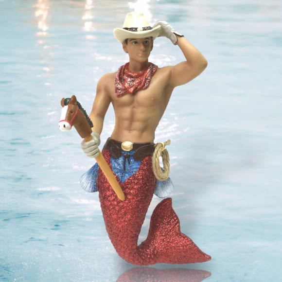 The Cowpoke Ornament by December Diamonds features a charming merman figurine with a red glittery tail, cowboy hat, red neckerchief, and white gloves. He holds a hobby horse and has a lasso at his side, set against the light blue water surface.
