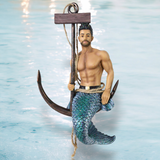 The Fresh Catch Ornament by December Diamonds showcases a merman with a muscular torso, blue-scaled tail, and short dark hair. Perched on a large anchor with a rope in hand against an oceanic backdrop, it beautifully captures the seas mystique.