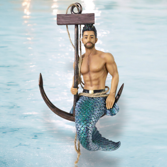 The Fresh Catch Ornament by December Diamonds showcases a merman with a muscular torso, blue-scaled tail, and short dark hair. Perched on a large anchor with a rope in hand against an oceanic backdrop, it beautifully captures the seas mystique.