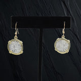 The Treasure Shoppe Inc.s Atocha Coin Gold Plated Earrings, featuring historic charm with gold accents, hang elegantly on hooks and are displayed on a black stand against a dark textured background.
