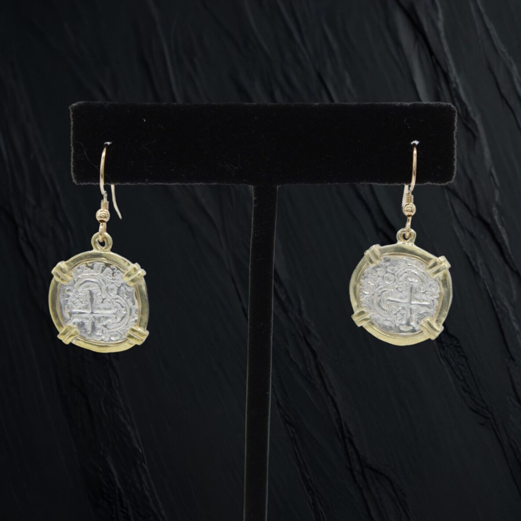 The Treasure Shoppe Inc.s Atocha Coin Gold Plated Earrings, featuring historic charm with gold accents, hang elegantly on hooks and are displayed on a black stand against a dark textured background.
