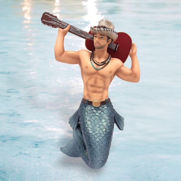The December Diamonds Guitar Man Ornament features a muscular merman wearing a hat and ornate necklace, holding a guitar over his shoulder. He has a blue-green fish tail and is depicted standing in water against a soft blue background.