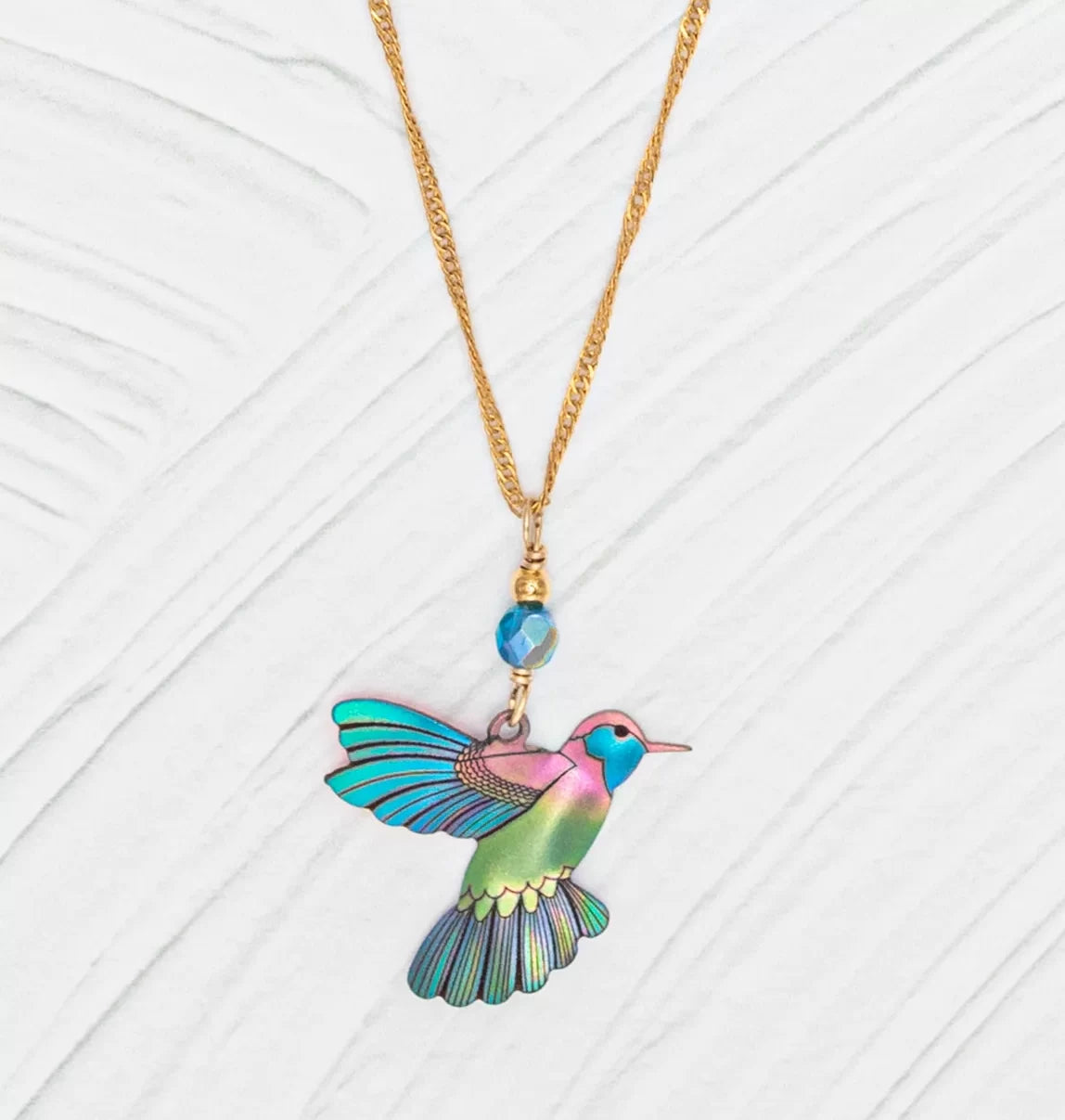 The Island Green Picaflor Necklace by Holly Yashi features a hand-painted niobium hummingbird in vibrant blues, greens, and purples. Its paired with an 18k gold-plated chain and a small blue bead, all on a white textured background.