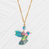 The Island Green Picaflor Necklace by Holly Yashi features a hand-painted niobium hummingbird in vibrant blues, greens, and purples. Its paired with an 18k gold-plated chain and a small blue bead, all on a white textured background.
