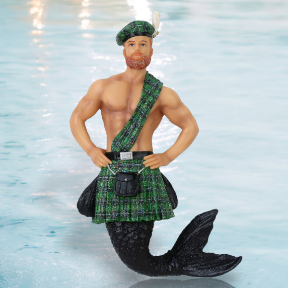 Keltic Kelly Ornament by December Diamonds features a merman with a black fish tail, dressed in a green and black plaid kilt, sash, and beret. He stands confidently with hands on hips against a light blue water backdrop, perfect for adding whimsy to your holiday decor.