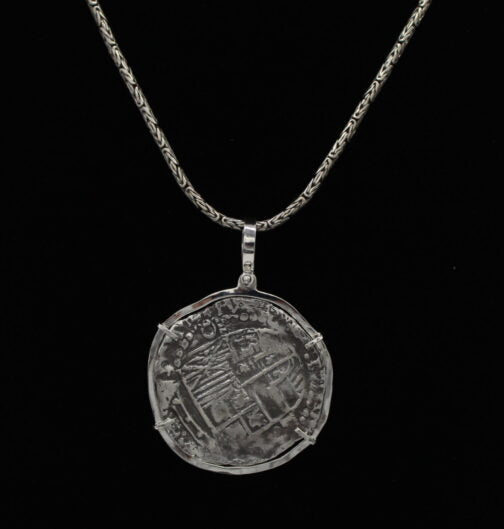 The Treasure Shoppe Inc. offers the Large Potosi Frame Coin, a pendant inspired by Atocha shipwreck coins. This intricate piece showcases a silver coin set in a sterling bezel against a solid black background, creating an exquisite design.