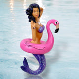 A mermaid with a sparkly purple tail and matching top poses in a pink flamingo float in a pool, embodying style and playfulness. Like an enchanting holiday decor piece from December Diamonds Miss Flamingo Ornament collection, she adds charm to any mermaid ornaments display.