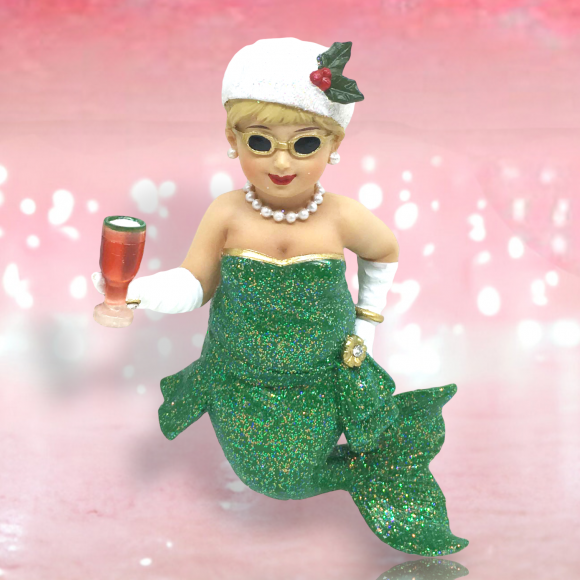 The Miss Holly Ornament by December Diamonds features a festive mermaid with blonde hair, glittery green tail, sunglasses, and a white holly-adorned hat. She holds a champagne glass and wears a pearl necklace against a pink, sparkly background. This hand-painted figure embodies holiday cheer.