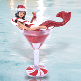 The December Diamonds Miss Peppermintini Ornament features a mermaid adorned with a red glittery tail and Santa hat, lounging in a cocktail glass filled with red liquid. Wrapped in a candy cane float, she holds a small cocktail against a soft, blurred blue-green background.