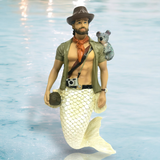 The Outback Ornament by December Diamonds features a hand-painted merman with a beard, hat, shirt, and orange scarf holding a camera and treasure chest while a koala sits on his shoulder. Perfect for holiday decor against a shimmering water surface backdrop.