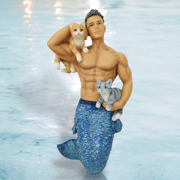 The Pet the Puss Ornament by December Diamonds features a muscular merman with a shiny blue tail, kneeling on a reflective surface. He playfully holds an orange cat on his shoulder and cradles a gray cat. This captivating piece is perfect for holiday decor or any mermaid collection.