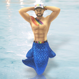 A bearded merman poses, hands behind his head, with December Diamonds vibrant Resist Ornament, including a white RESIST cap, rainbow bracelet and chest paint, a shimmering blue tail, and a rainbow belt highlighting his muscular torso against the watery backdrop.