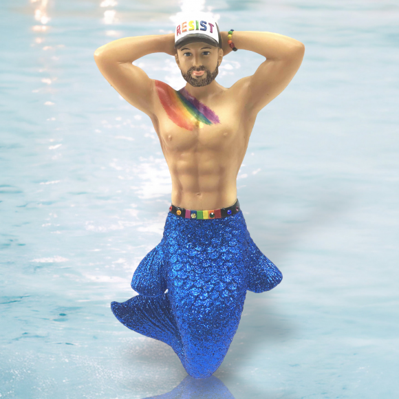 A bearded merman poses, hands behind his head, with December Diamonds vibrant Resist Ornament, including a white RESIST cap, rainbow bracelet and chest paint, a shimmering blue tail, and a rainbow belt highlighting his muscular torso against the watery backdrop.