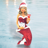 The Santa Baby II Ornament by December Diamonds is a mermaid in a festive Santa outfit with a red and white hat, holding a silver-wrapped gift and red martini glass. Her shimmering red-scaled tail stands out against the soft, blurred watery background.