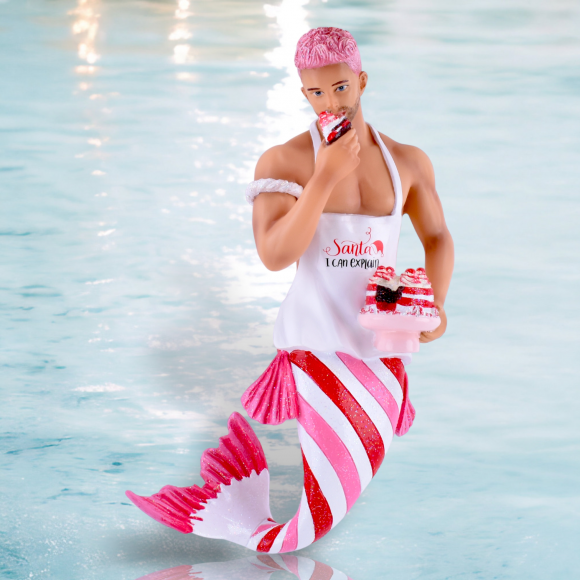 The Santas Taster Ornament by December Diamonds features a merman with pink hair, striped tail, and a Santa I Can Explain apron as he enjoys cupcakes. Its whimsical design is perfect for holiday decor against a watery backdrop.