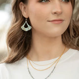 A woman in a white shirt and leaf-shaped earrings exudes elegance. Her long brown hair cascades over two necklaces: Holly Yashis Shelby Sage and Gold Necklace, and another featuring an exquisite seashell pendant, with a softly blurred background adding mystery to her serene presence.