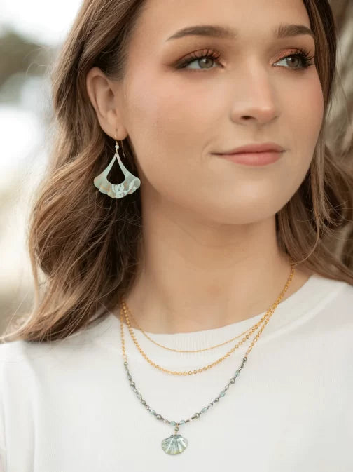 A woman in a white shirt and leaf-shaped earrings exudes elegance. Her long brown hair cascades over two necklaces: Holly Yashis Shelby Sage and Gold Necklace, and another featuring an exquisite seashell pendant, with a softly blurred background adding mystery to her serene presence.