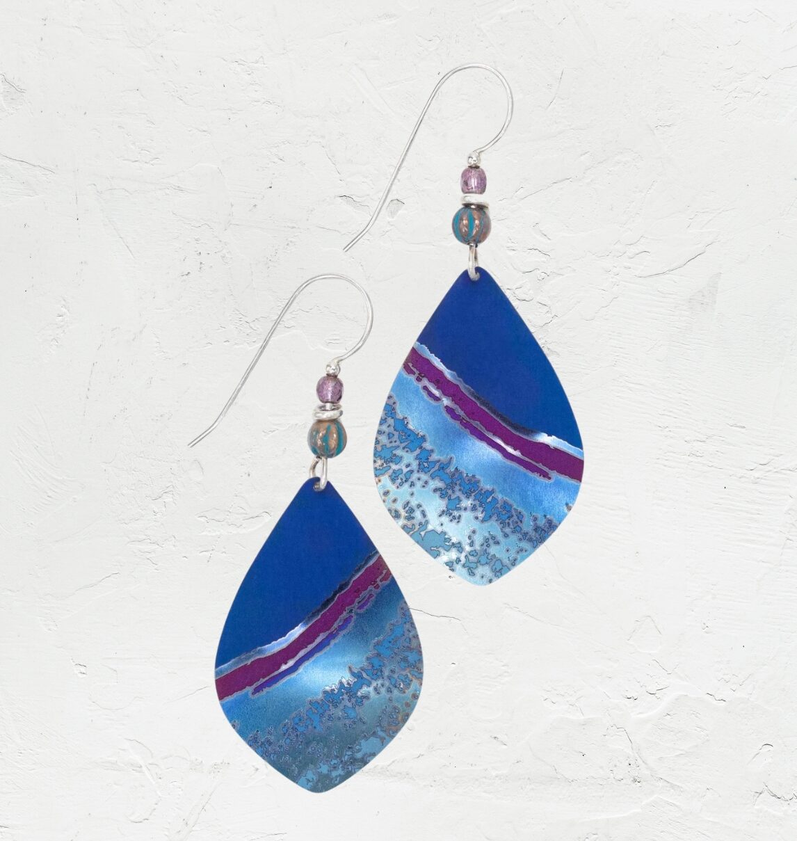 The Shorebreak Blue Depths Earrings from Holly Yashi feature teardrop jewels in a stunning blue, purple, and silver gradient. They dangle gracefully from hooks embellished with small round beads, set on a textured white background—an ideal treasure for beach enthusiasts.