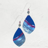 The Shorebreak Blue Depths Earrings from Holly Yashi feature teardrop jewels in a stunning blue, purple, and silver gradient. They dangle gracefully from hooks embellished with small round beads, set on a textured white background—an ideal treasure for beach enthusiasts.