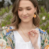 A woman with long hair softly smiles, holding a starfish pendant. She wears a white blouse, a colorful floral scarf, and Shorebreak Blue Depths Earrings by Holly Yashi that shimmer like a mermaids tear. The blurred greenery in the background provides an ideal setting for beach lovers.