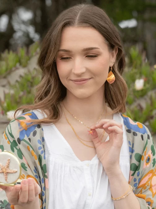 A woman with long hair softly smiles, holding a starfish pendant. She wears a white blouse, a colorful floral scarf, and Shorebreak Blue Depths Earrings by Holly Yashi that shimmer like a mermaids tear. The blurred greenery in the background provides an ideal setting for beach lovers.