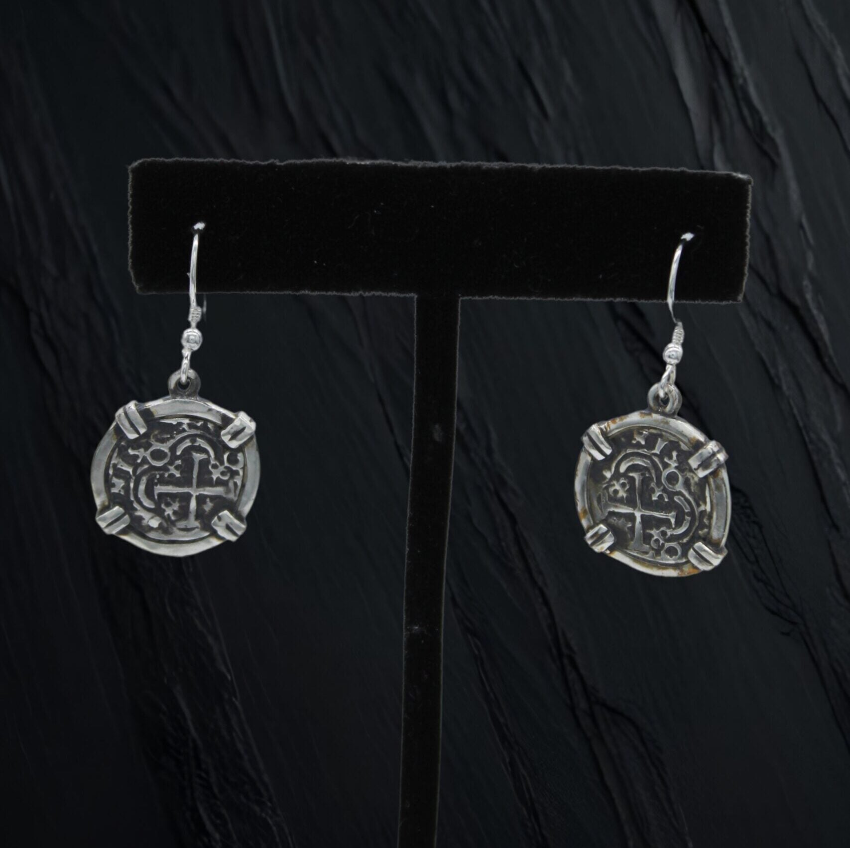 The Treasure Shoppe Inc.s Small Wrapped Coin Earrings feature Atocha coin designs crafted from sterling silver. Displayed on a black stand, they glimmer with intricate, antique-inspired details perfect for fans of unique, timeless accessories.