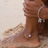 A close-up shows a sandy foot wearing Shanores Swarovski Crystals White Sand Dollar Anklet-Ocean Jewelry with a starfish charm. As they adjust it on the beach, their hand reveals matching bracelet and rings sparkling in the sunlight.