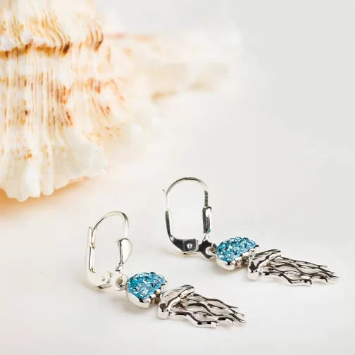 These sterling silver earrings from Shanore feature intricate leaf designs adorned with blue gemstones. The Swarovski Crystals Aqua Jellyfish Earrings from their Ocean Jewelry line, shine elegantly against a light surface, with a blurred seashell backdrop reminiscent of jellyfish glistening underwater.