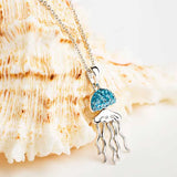 The Shanore Swarovski Crystals Aqua Jellyfish Necklace features a pendant resembling a jellyfish with a blue gemstone body and sterling silver tentacles. It hangs on a silver chain against a cream and brown seashell background.