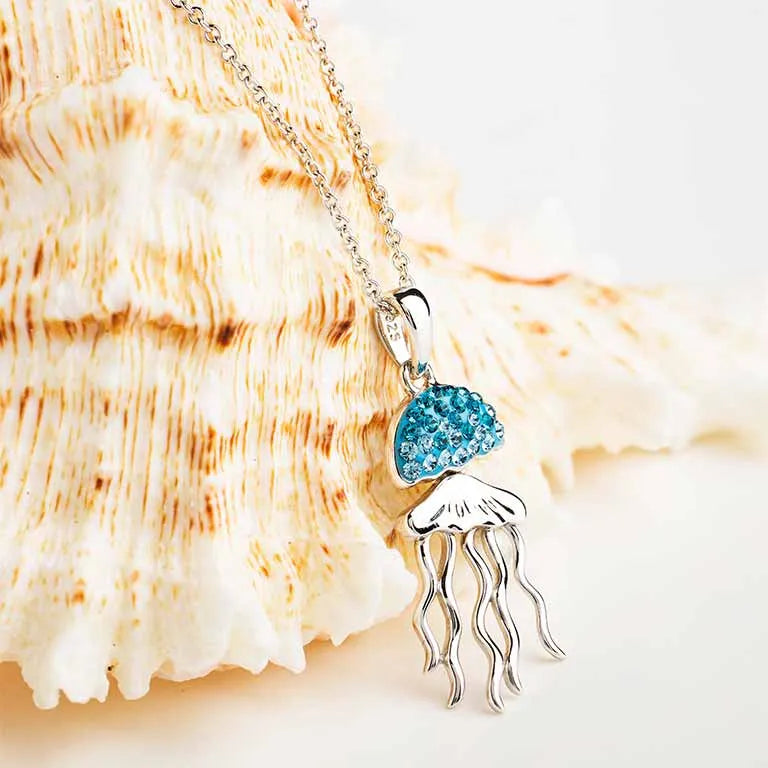 The Shanore Swarovski Crystals Aqua Jellyfish Necklace features a pendant resembling a jellyfish with a blue gemstone body and sterling silver tentacles. It hangs on a silver chain against a cream and brown seashell background.