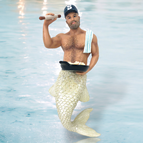 The Sticky Buns Ornament by December Diamonds features a muscular merman with a shimmering tail, holding a rolling pin and pan of baked goods. A towel drapes over his shoulder, and he wears an S cap. This hand-painted ornament, set against a watery backdrop, is perfect for holiday decor.