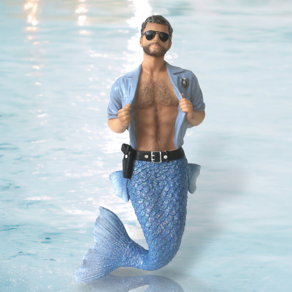 The Strip Search Ornament by December Diamonds features a hand-painted merman with a blue sparkly tail, wearing a partially open light blue shirt and sunglasses against a bright blue water background. Its ideal for unique holiday decor enthusiasts or Merman and Mermaid ornament collectors.
