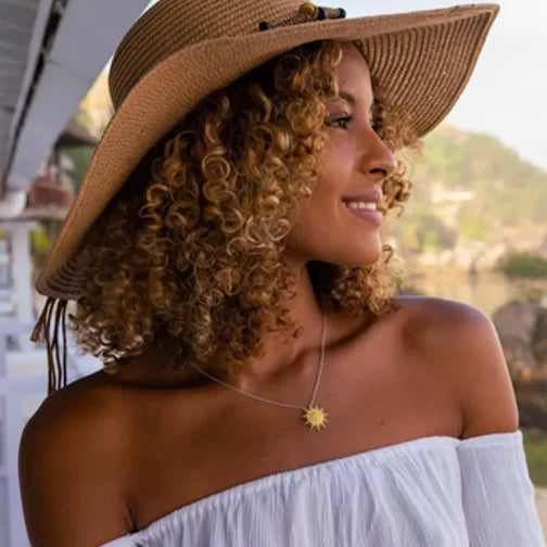 A curly-haired woman in a wide-brimmed straw hat and an off-the-shoulder white top looks left, flaunting the Shanore Ocean Jewelry Sterling Silver and Swarovski Crystals Sunburst Necklace. The blurred background suggests an outdoor setting.