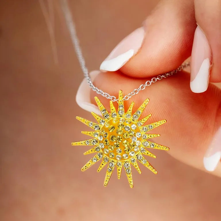 A manicured hand showcases the Shanore Sterling Silver and Swarovski Crystals Sunburst Necklace from Ocean Jewelry, featuring a captivating sunburst pendant adorned with sparkling crystals.