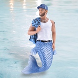 The December Diamonds Trucker Ornament is a captivating merman featuring a blue-scaled tail set against a watery backdrop. He wears a white tank top, blue cap, and has a checkered towel over his shoulder, complete with tattoos and belt—a confident figure ideal for holiday decor charm.