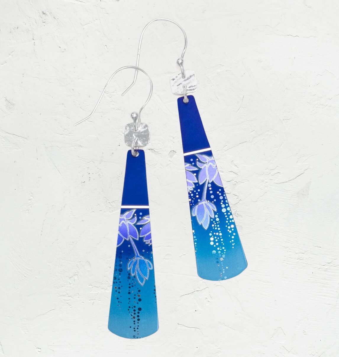 The Midnight Blue Tula Earrings by Holly Yashi Jewelry feature a blue gradient with silver hooks and floral designs. Adorned with decorative dots, theyre hypo-allergenic and set against a textured surface, enhanced by elegant 18k gold-filled accents.