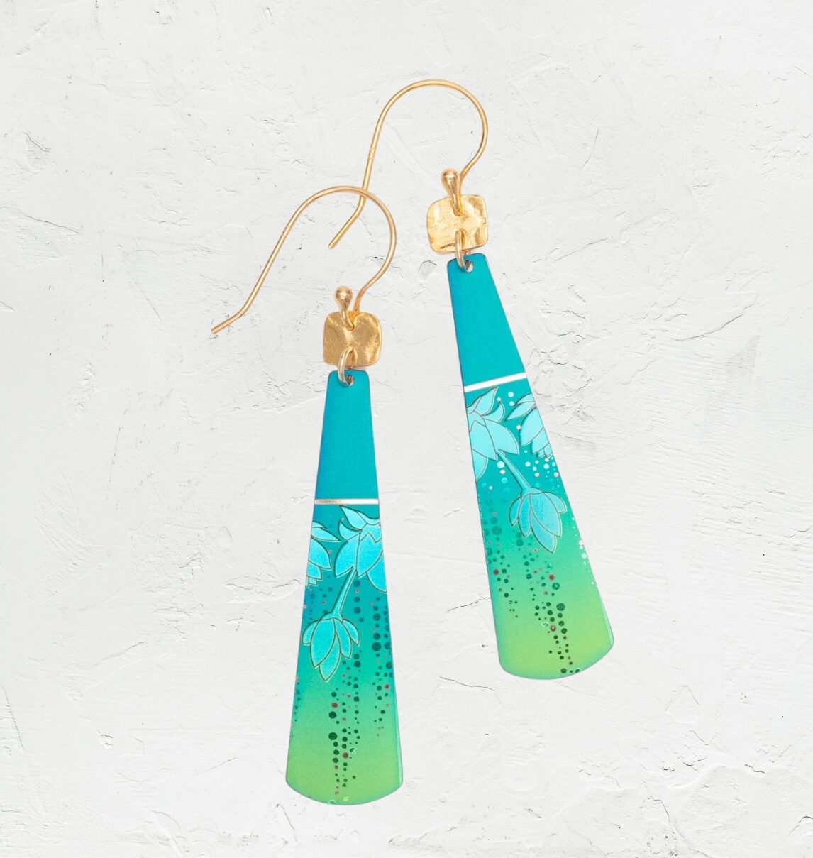 Presenting the Turquoise Tula Earrings by Holly Yashi: exquisite teardrop earrings with a teal-to-lime gradient, floral pattern, and gold hooks. Made from hypo-allergenic niobium, they elegantly hang against a textured white backdrop.