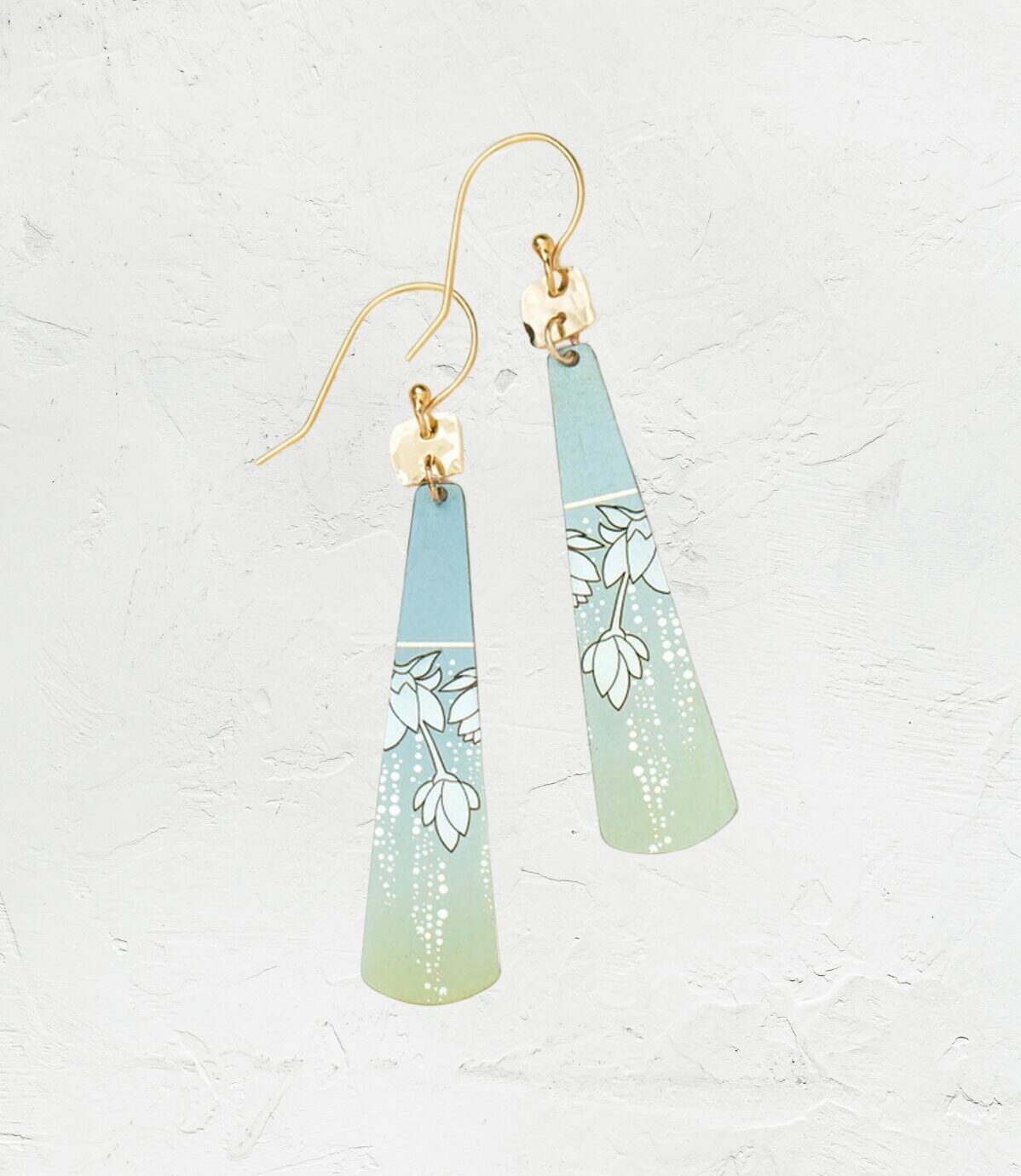 The Sage Tula Earrings by Holly Yashi feature hypo-allergenic, niobium-crafted triangular pieces with a blue-to-green gradient and floral designs, finished with gold hooks and set on a textured white background.