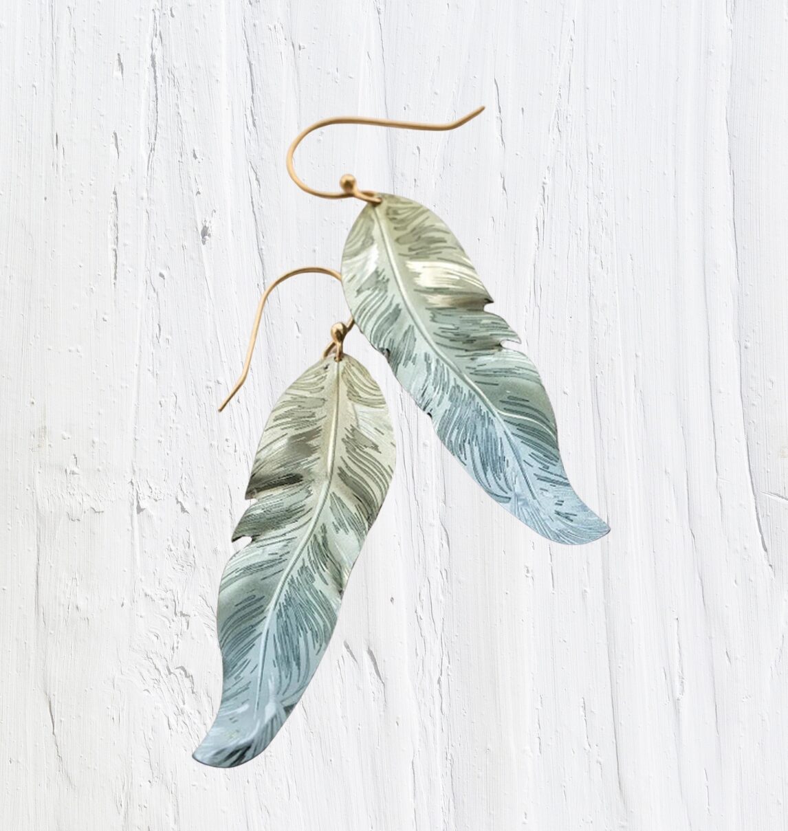 The Free Spirit Feather Sage Earrings by Holly Yashi, made from niobium, gracefully dangle with a metallic sheen against a white textured background. With Bohemian flair, they showcase gradient feathers transitioning from silver to light blue.