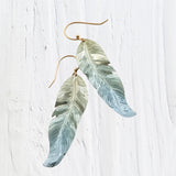 The Free Spirit Feather Sage Earrings by Holly Yashi, made from niobium, gracefully dangle with a metallic sheen against a white textured background. With Bohemian flair, they showcase gradient feathers transitioning from silver to light blue.