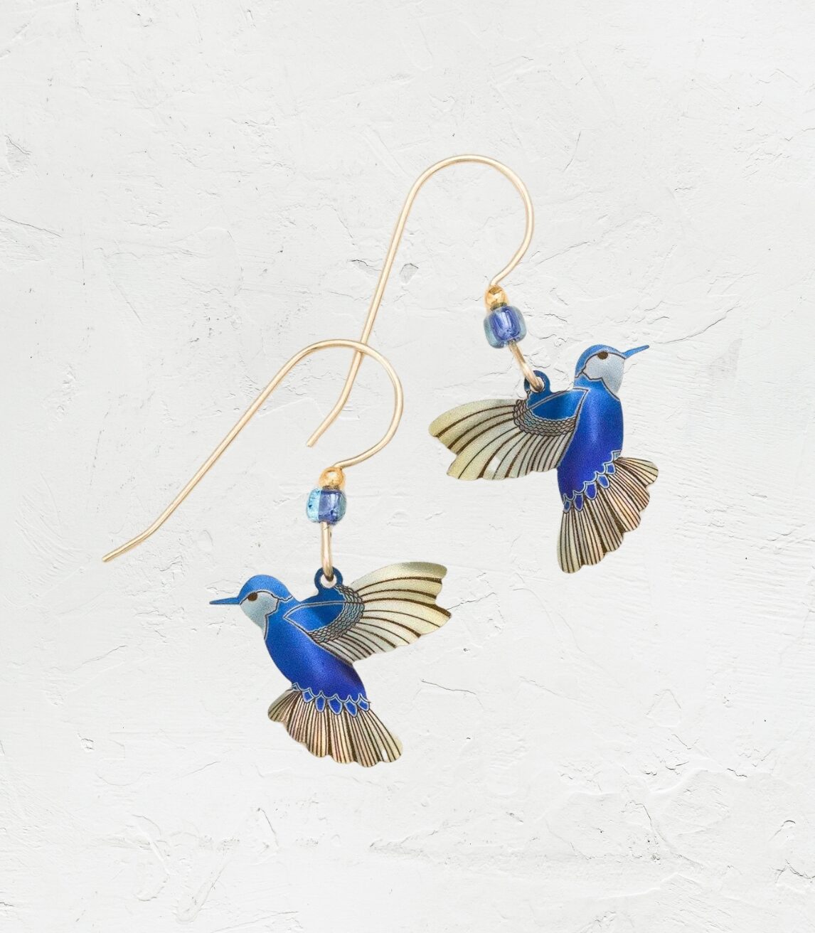 Discover the enchanting Blue Picaflor Earrings by Holly Yashi: hummingbird-shaped earrings with 18k gold-filled wings and tails, adorned with small blue beads on a textured white surface for a delicate jewelry collection addition.