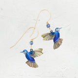 Discover the enchanting Blue Picaflor Earrings by Holly Yashi: hummingbird-shaped earrings with 18k gold-filled wings and tails, adorned with small blue beads on a textured white surface for a delicate jewelry collection addition.