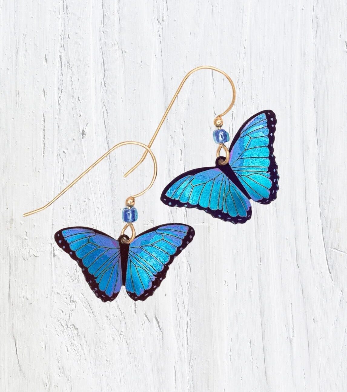 The Blue Bella Butterfly Earrings by Holly Yashi feature vibrant blue wings with black edges, suspended from gold hooks. Each handcrafted piece includes a small blue bead near the hook and is made of lightweight niobium against a textured white surface.