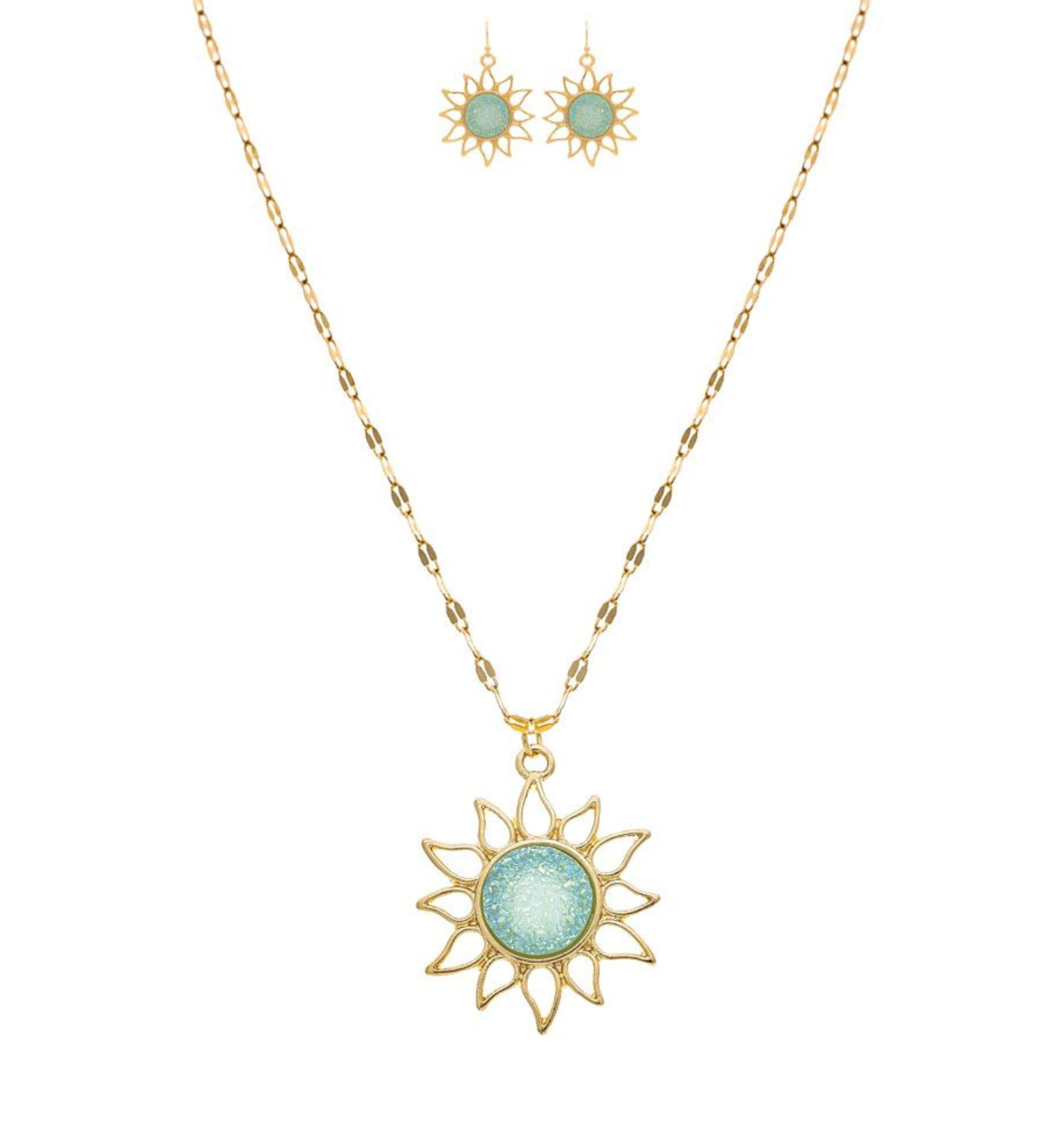 The A-Train Gold Blue Druzy Sun Necklace Set features a sun-shaped pendant with a round, light blue center and matching earrings, crafted from premium jeweler’s metals and shown on a white background.