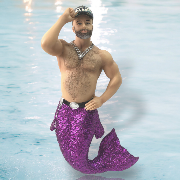 The December Diamonds Whos Your Daddy Ornament features a hand-painted, bearded merman with a muscular build and glittery purple tail, saluting with one hand. Wearing a starry cap and set against a watery light blue background, it makes an ideal piece for holiday decor.