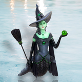 The Wicked Ornament by December Diamonds features a whimsical mermaid with a green complexion and black dress, adorned with a witch hat and broom. She floats on water holding a glowing green orb against a light blue background—a perfect holiday decor piece.
