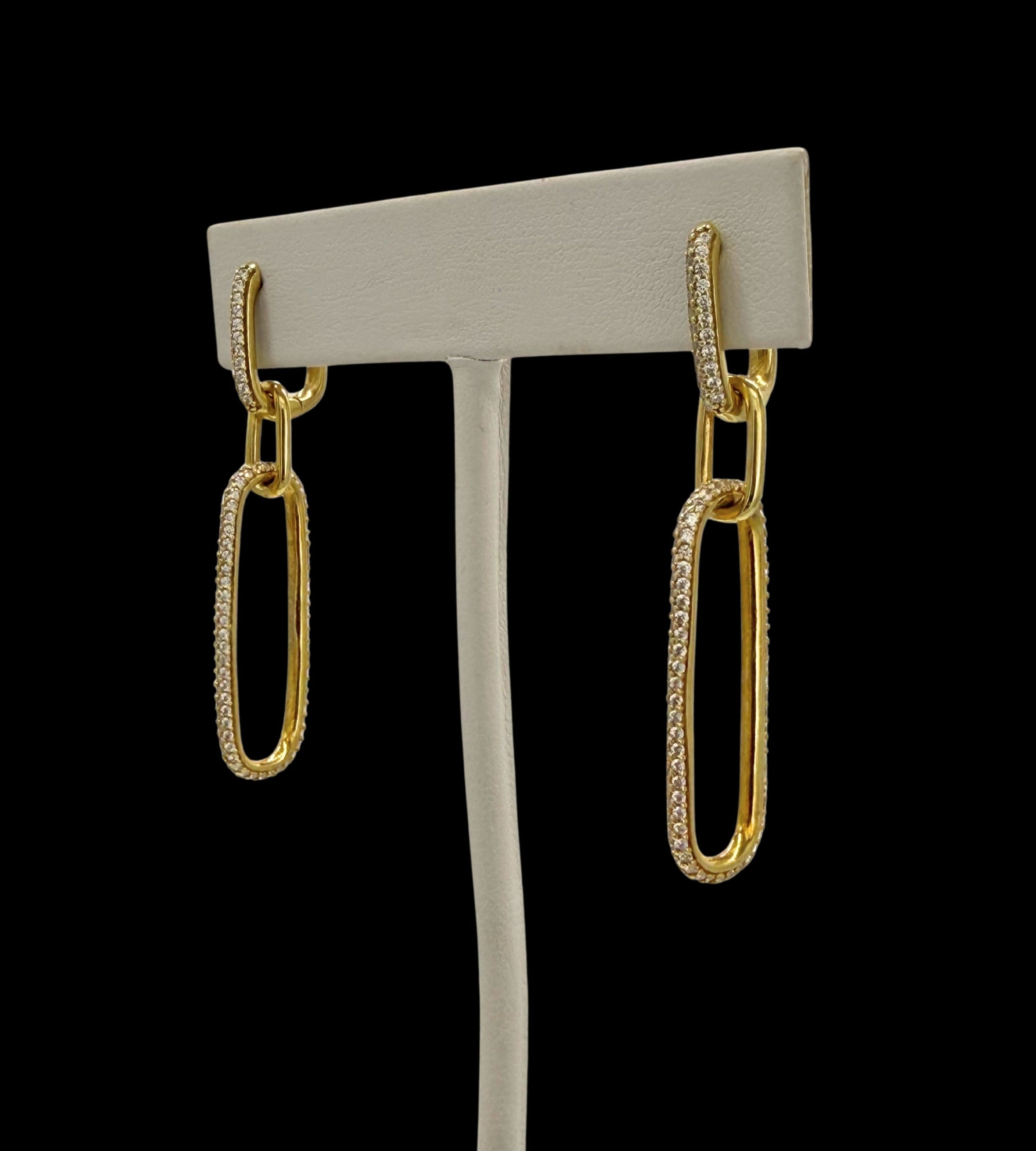 The Be-Je Designs Gold Pave CZ Double Link Earrings feature handcrafted 14kt gold elongated oval links, some adorned with sparkling cubic zirconia, displayed on a simple gray stand against a black background.