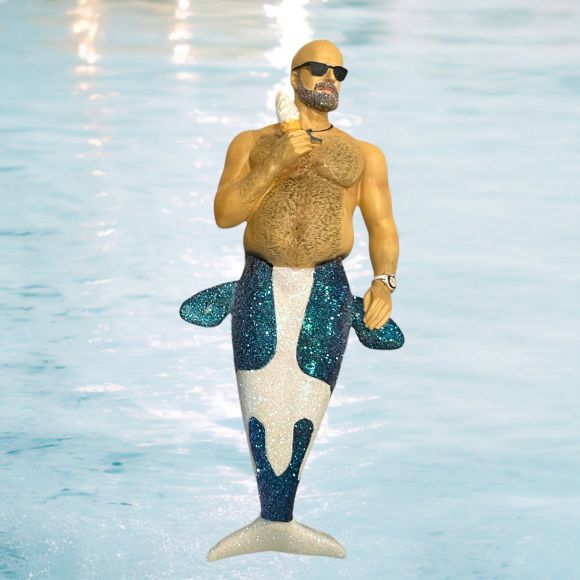 The December Diamonds Beached Whale/Blue Tail Ornament features a muscular, bearded figure with sunglasses and a sparkling orca tail, holding a banana, floating whimsically against a light blue water background.
