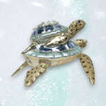 The December Diamonds ORN: Blue Turtle w/Baby on Back features a sparkling blue and silver shell with beads and jewels, displayed on a shimmering surface.